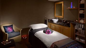 A spa room with two beds, a massage table, a sink, and a shelf with towels and toiletries.