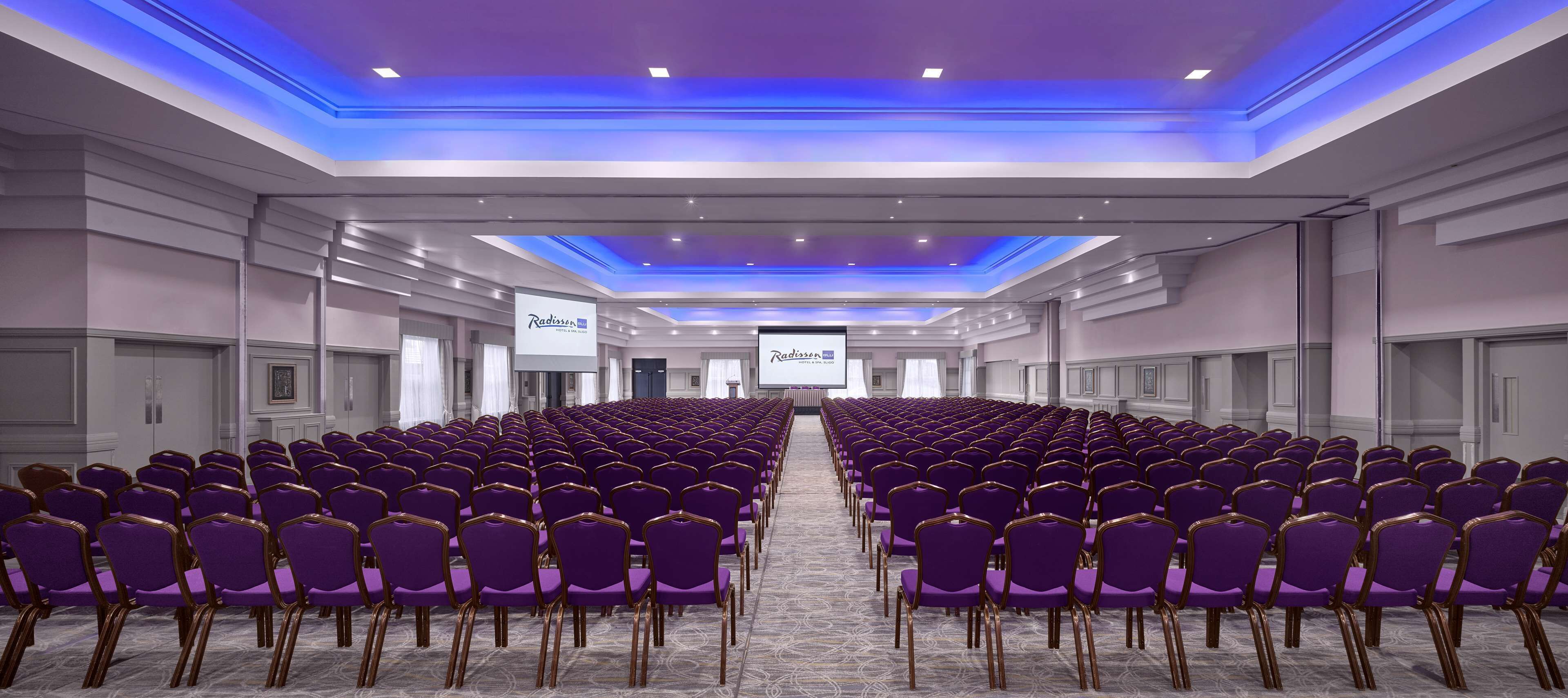 A conference room with rows of purple chairs and a large screen at the front.