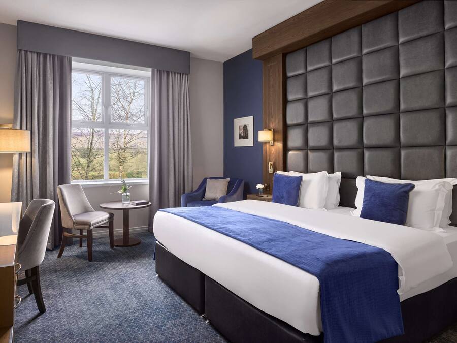 Una habitación de hotel con una cama grande, un escritorio, una silla, una mesita y un ventanal con cortinas.
