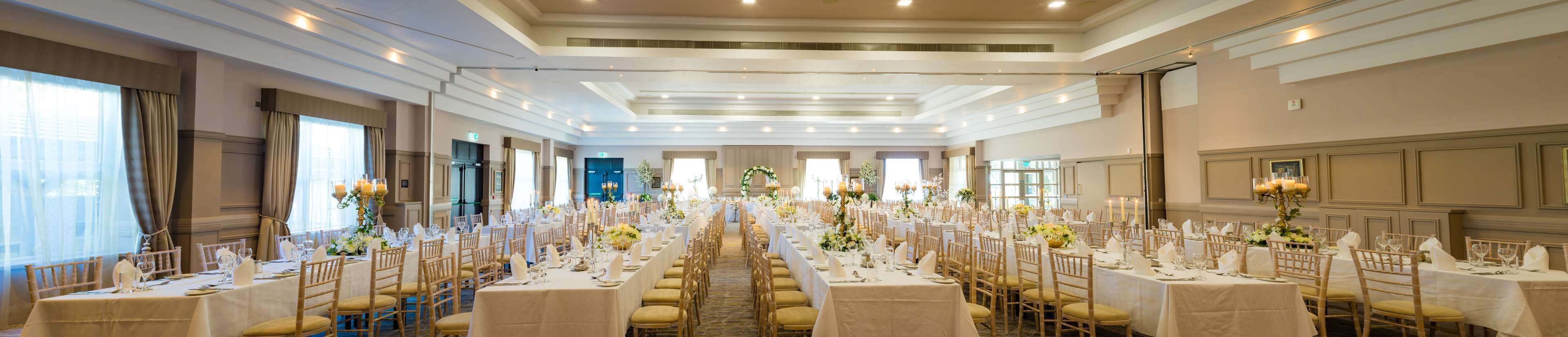 A banquet hall with long tables set for a formal event, featuring chandeliers and floral centerpieces.