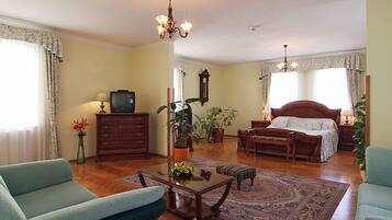 A spacious living room with a sofa, a coffee table, a television, and a bed with a wooden headboard. There are potted plants and a chandelier.