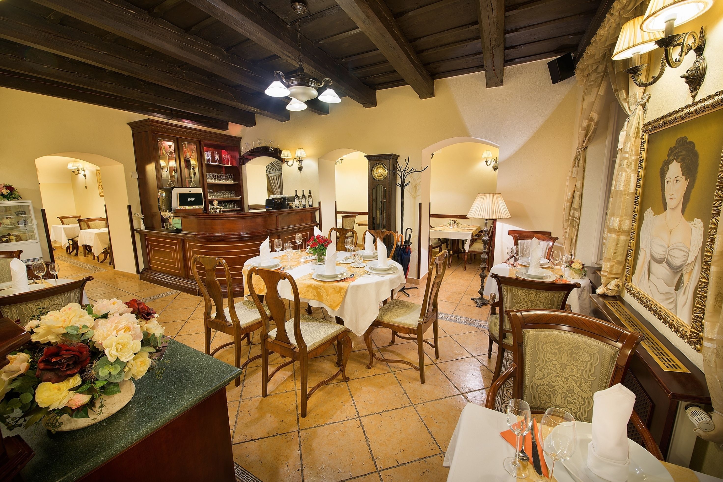 A dining area with wooden chairs and tables, a bar area with a wine rack, and a large framed portrait on the wall.