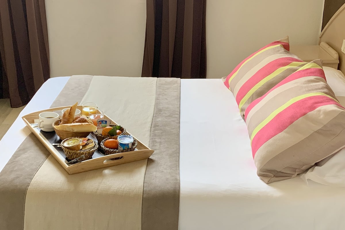 Una cama bien tendida con un almohadón a rayas, una bandeja de desayuno con pan, fruta y yogur, y una ventana con cortinas.