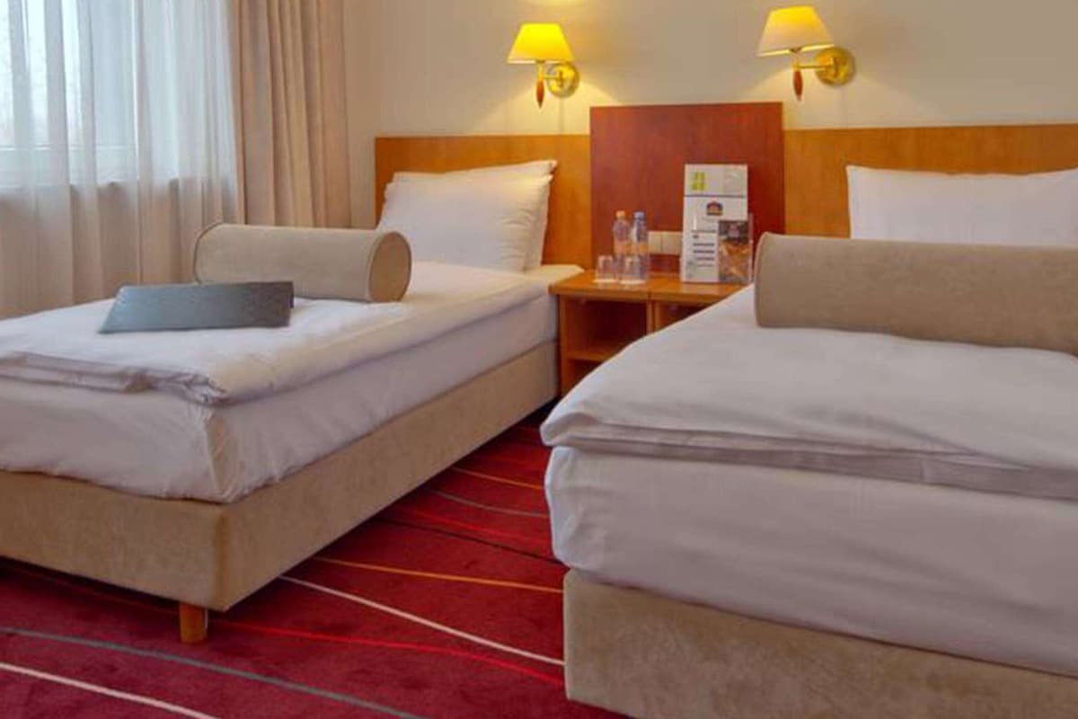 A hotel room with two single beds, a wooden headboard, a nightstand with a lamp, and a red carpet.