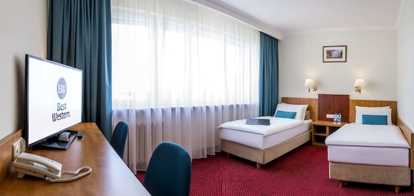 Habitación de hotel con una cama, un escritorio con monitor de computadora, un teléfono y una ventana con cortinas.