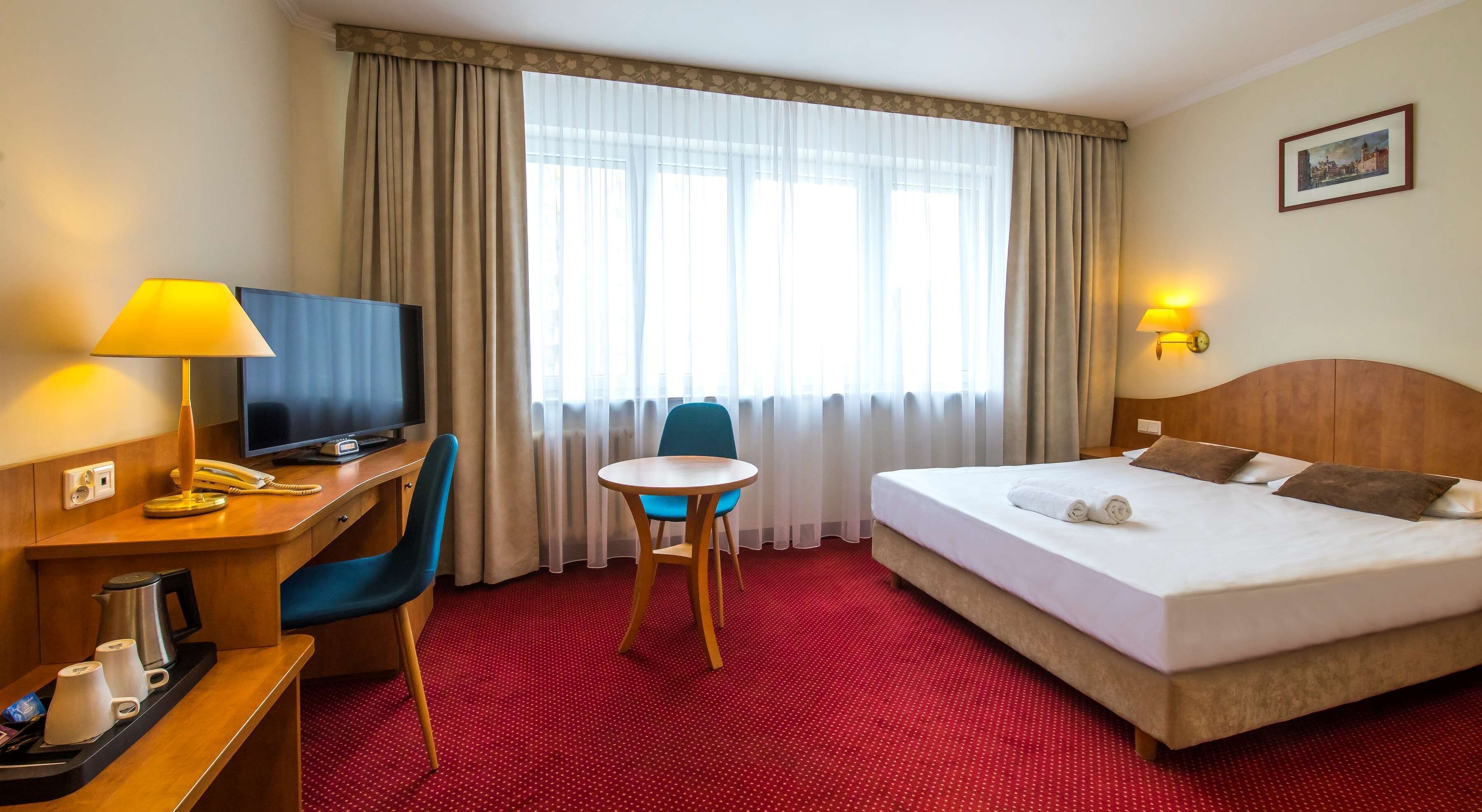 Ein Hotelzimmer mit Bett, Schreibtisch mit Fernseher, Sessel, kleinem Tisch und Fenster mit Vorhängen.