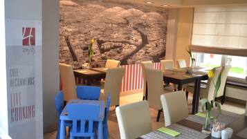 Un restaurant avec des chaises bleues et beiges, une grande fresque murale et un panneau indiquant que les plats sont préparés devant les clients.