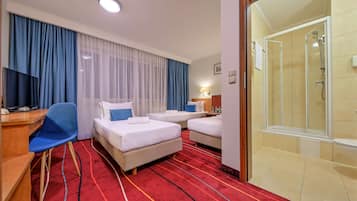 Hotellihuone, jossa on kaksi sänkyä, työpöytä, tuoli, televisio ja suihkutila.