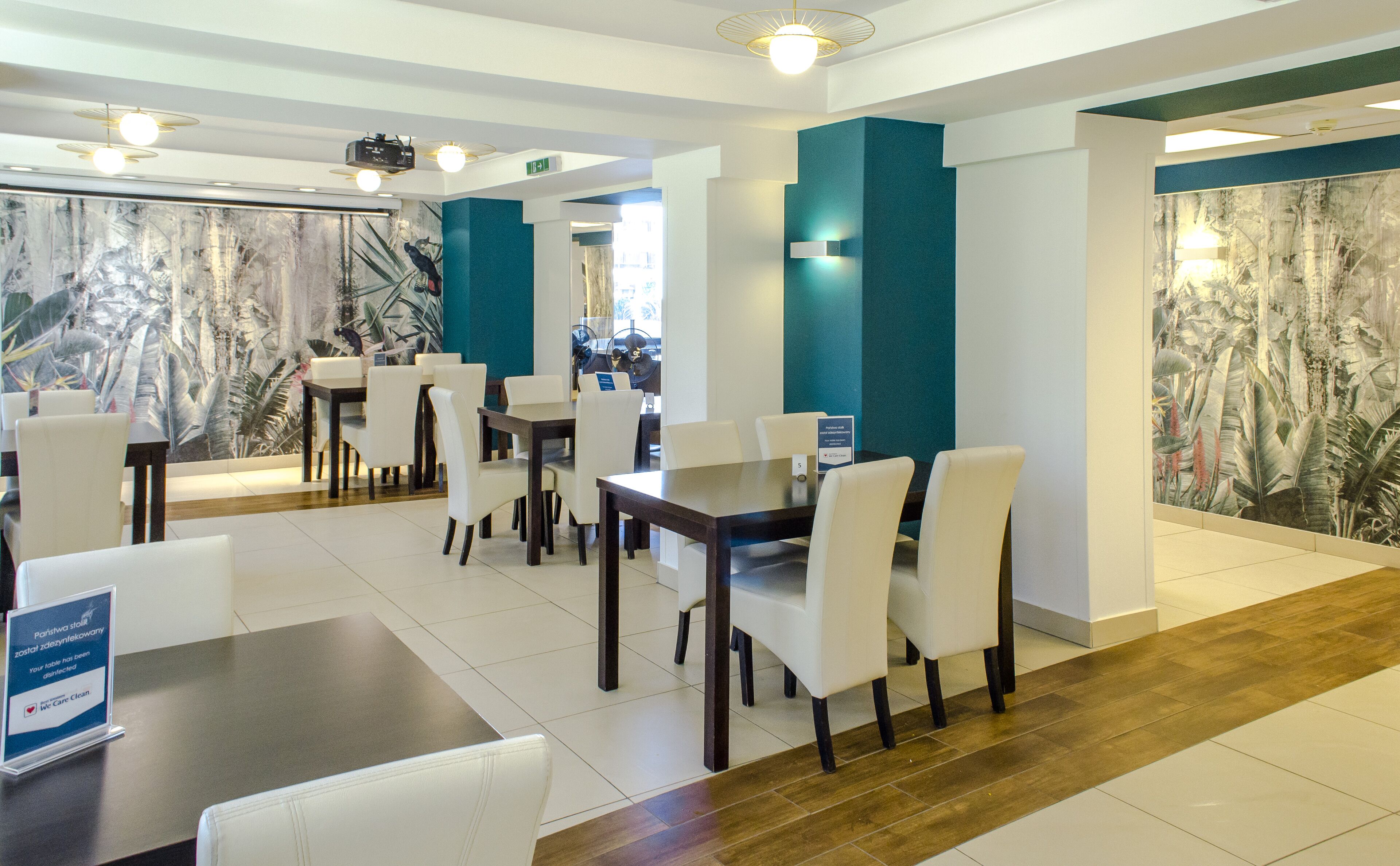 A modern dining area with white chairs and dark wooden tables, teal accent walls, and a large mural of tropical foliage.