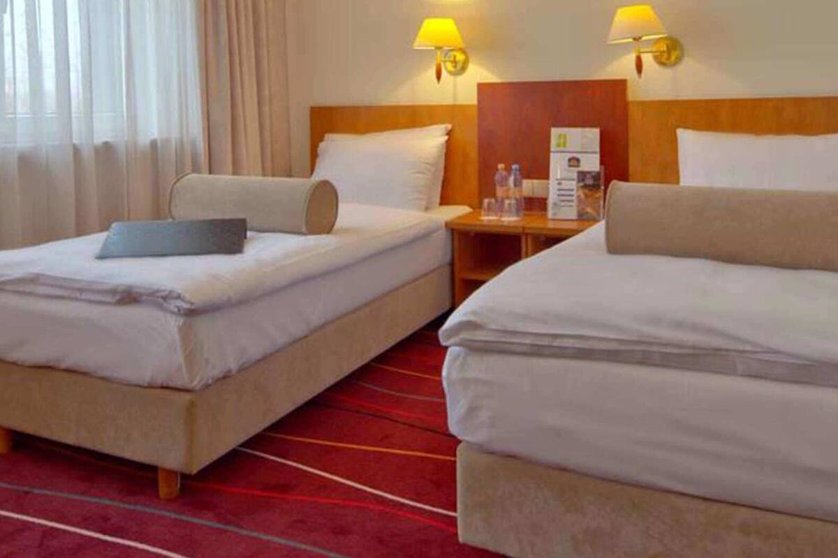 A hotel room with two single beds, a wooden headboard, a nightstand with a lamp, and a red carpet.
