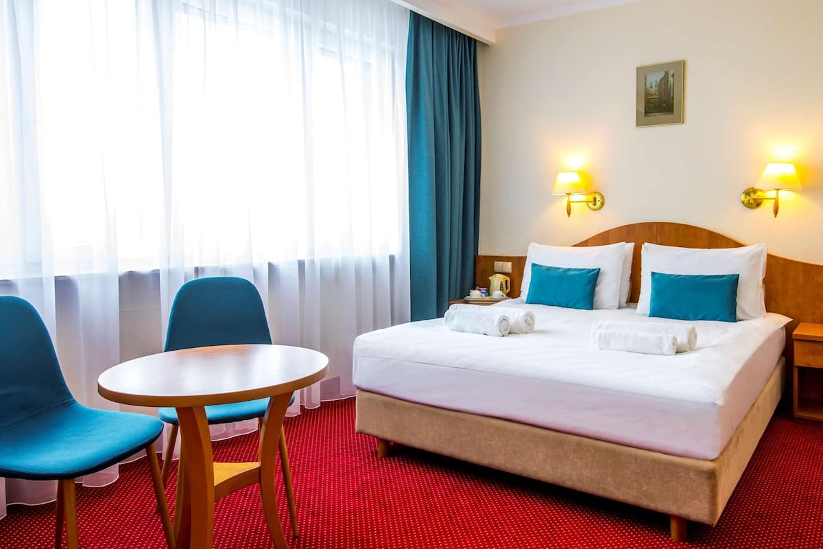 A hotel room with a bed, two chairs, a small table, a nightstand, and a window with curtains.