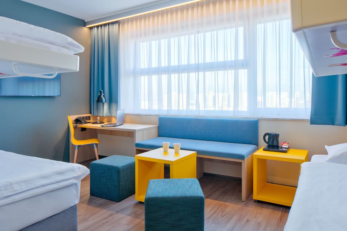 A modern hotel room with a bed, a desk, a blue sofa, and a small table with a kettle.