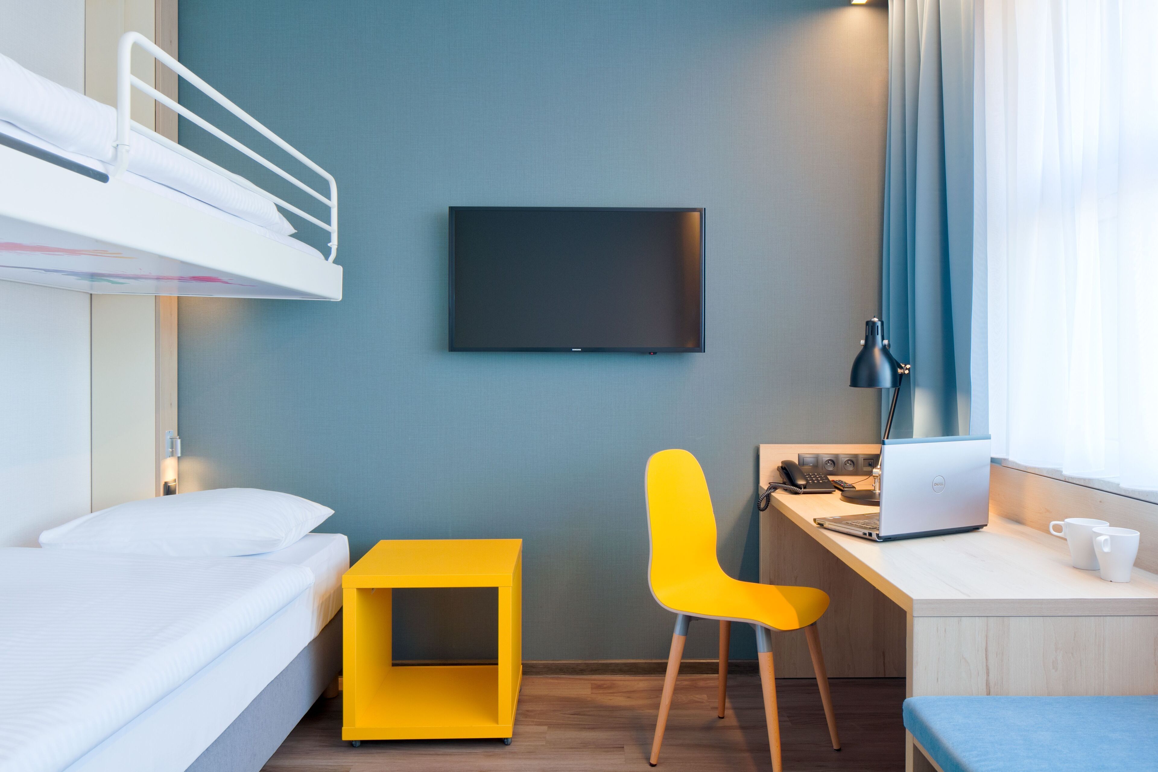 Een kamer met een stapelbed, een bureau met een laptop, een gele stoel en een televisie aan de muur.