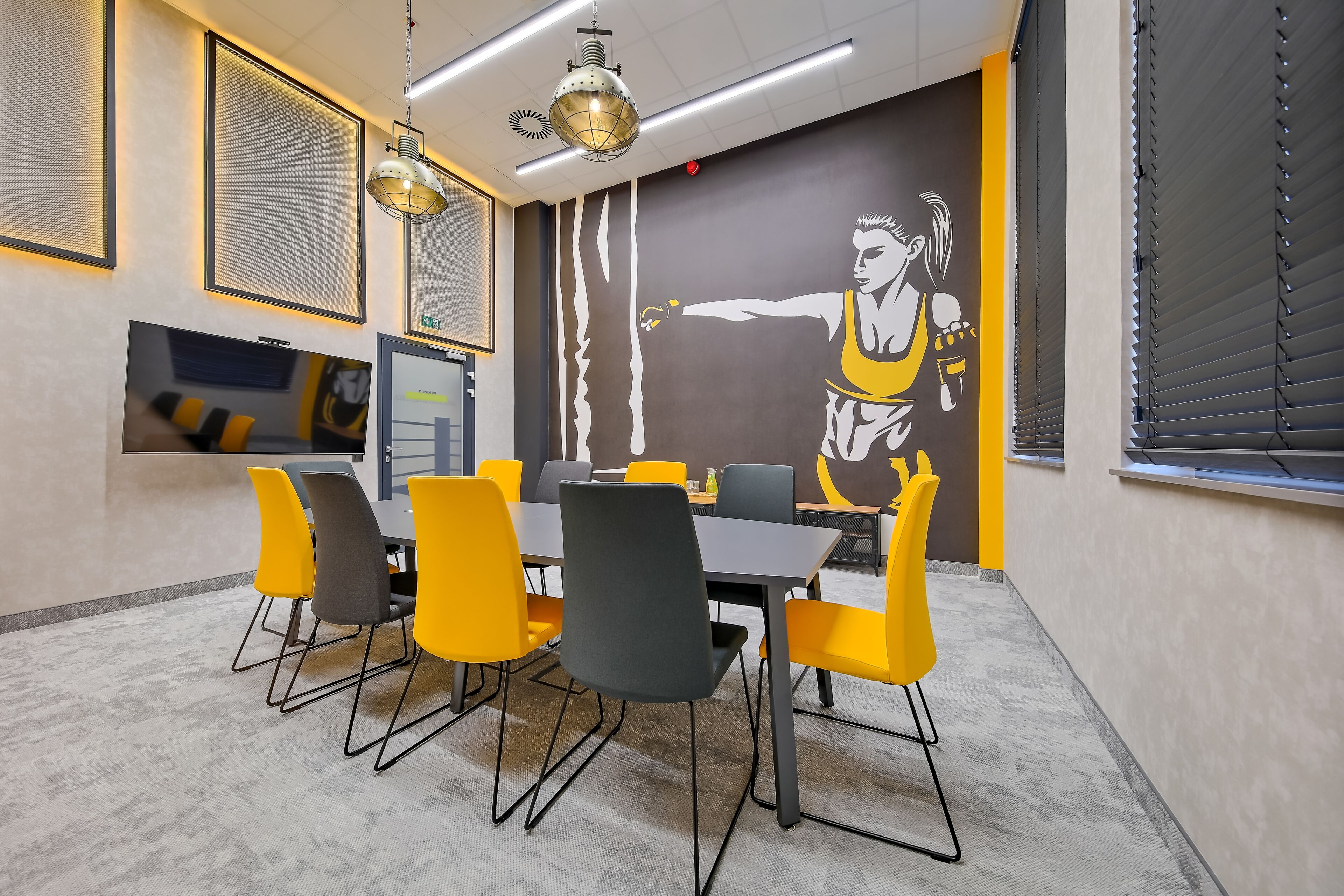 Un salón de conferencias con una gran mesa, sillas amarillas y grises, un mural y luminarias modernas.