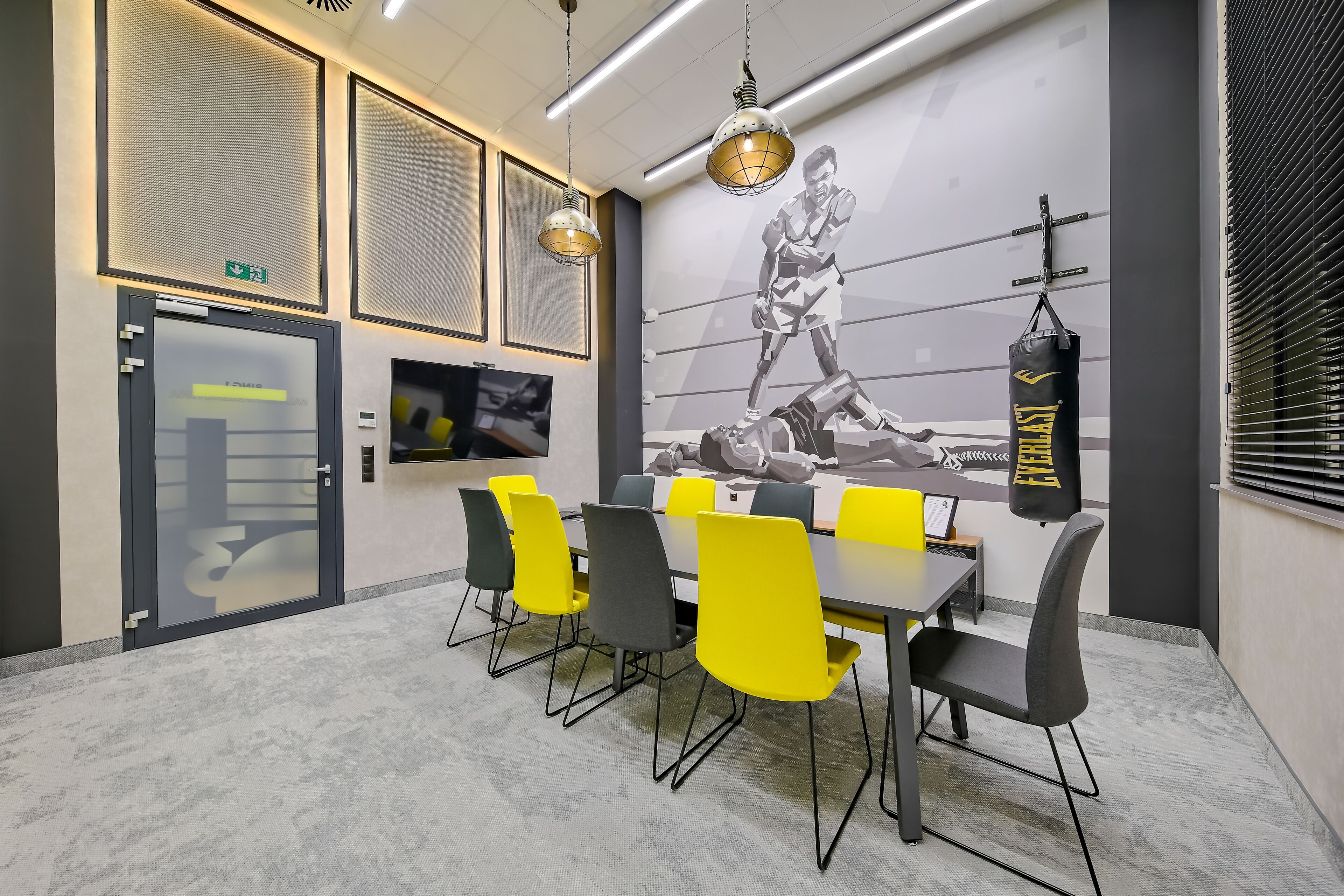 Un salón de conferencias con una gran mesa, sillas amarillas y grises, un saco de boxeo y un mural de un boxeador.