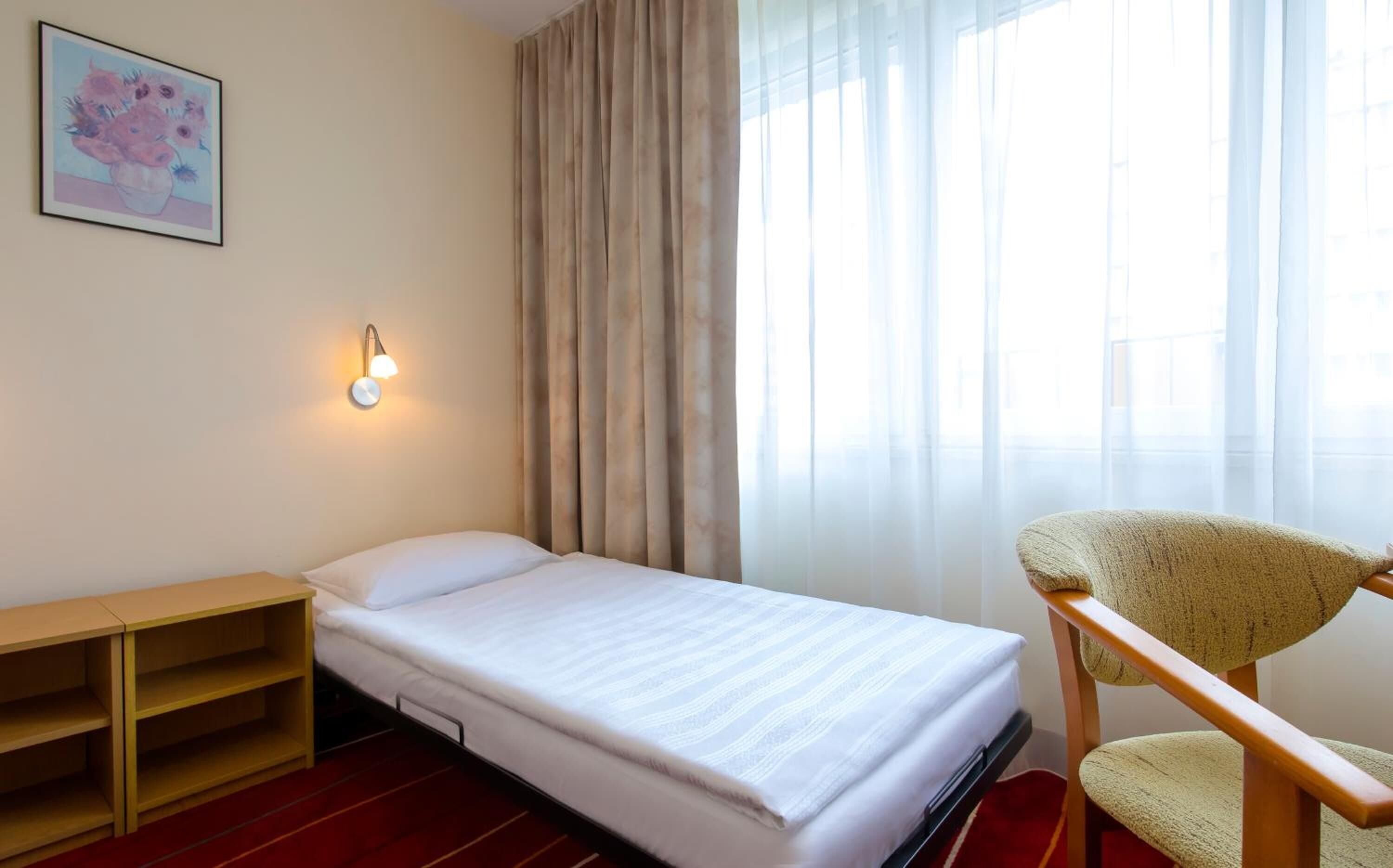 A compact hotel room with a single bed, a small wooden desk, a chair, and a large window with sheer curtains.