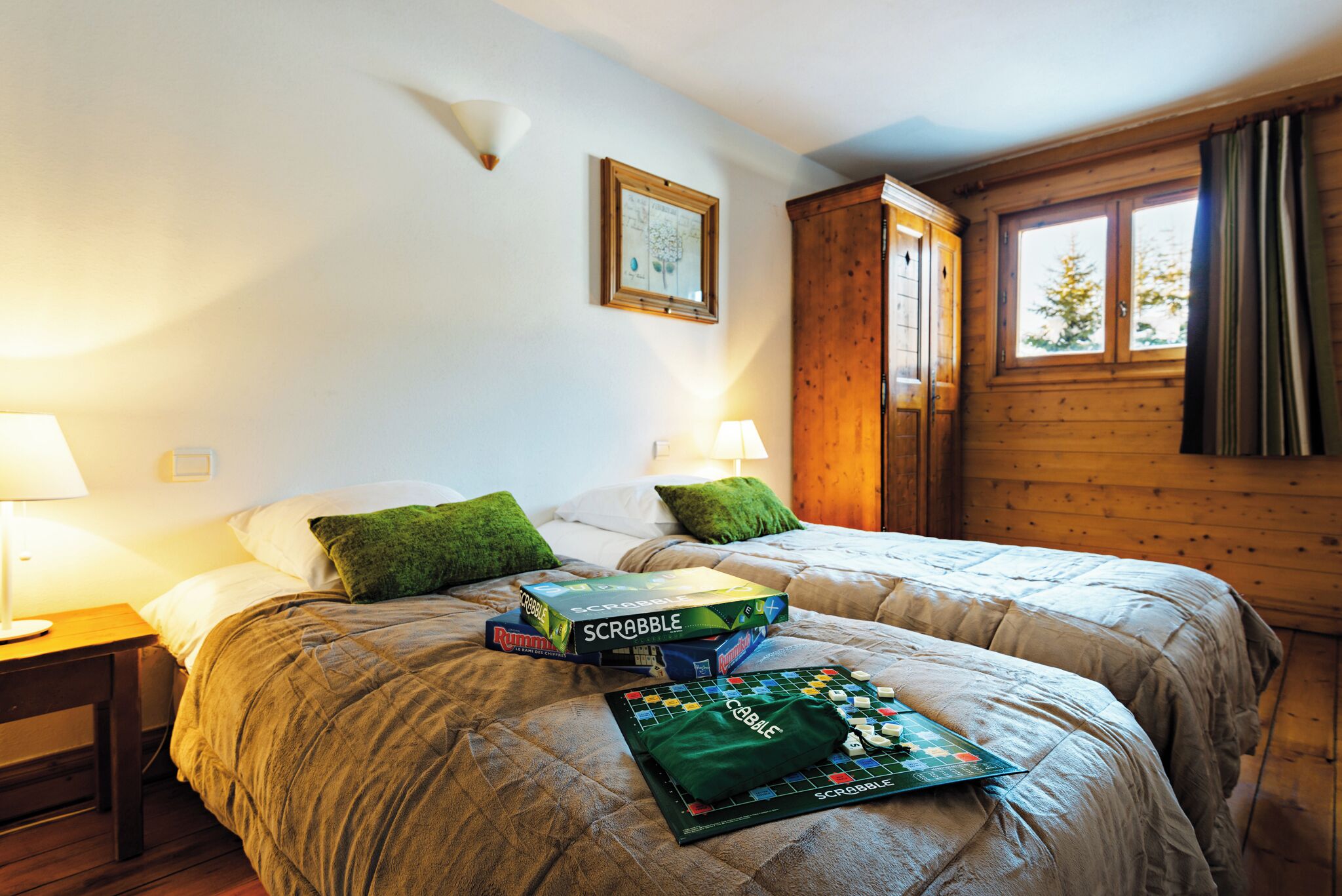 Une chambre avec deux lits, une armoire en bois, un tableau encadré au mur et des boîtes de jeu de Scrabble posées sur les lits.