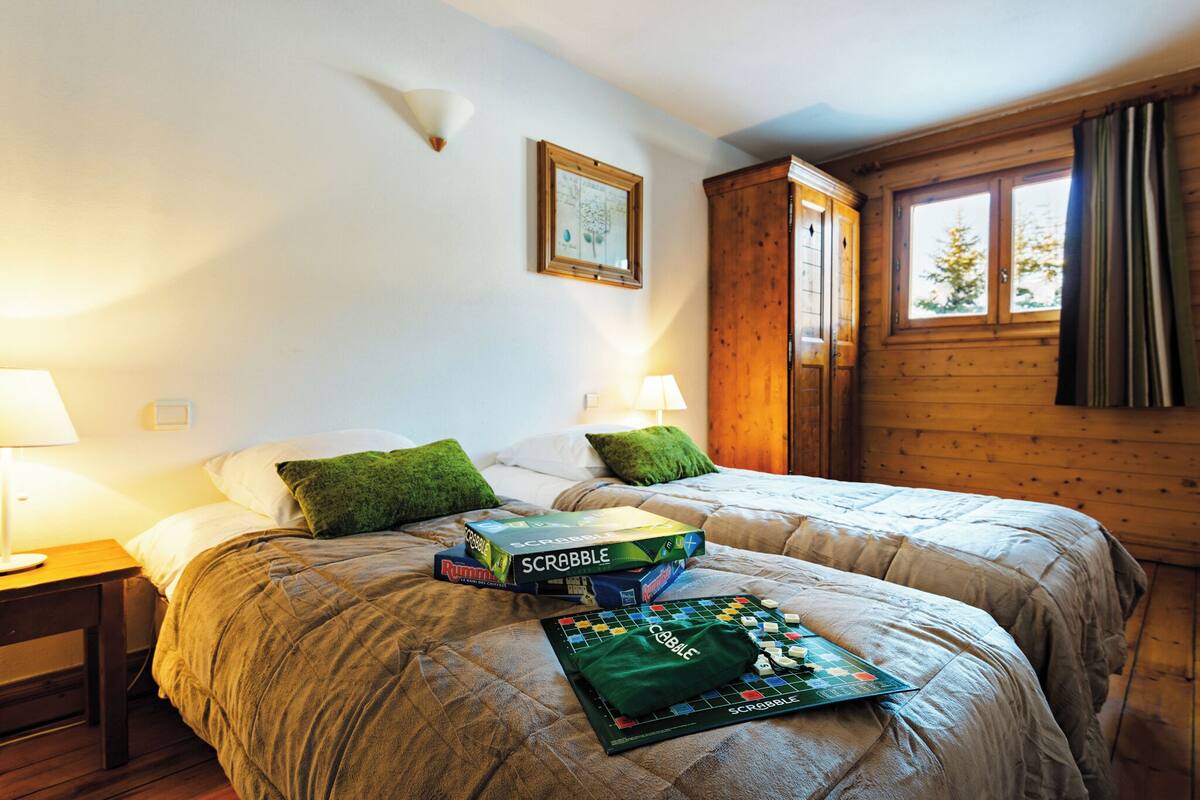 Ein Schlafzimmer mit zwei Betten, einem hölzernen Kleiderschrank, einem gerahmten Bild an der Wand und Scrabble-Spielkästen auf den Betten.