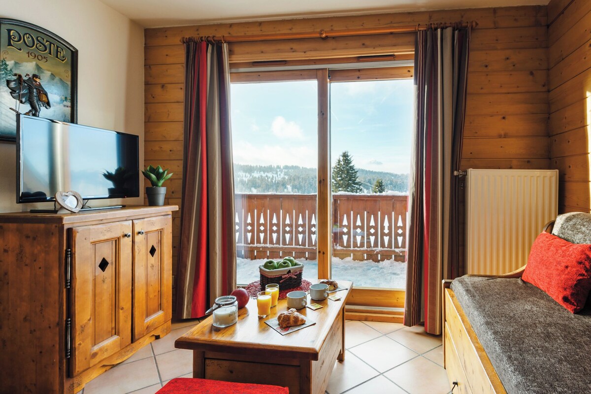 A wooden cabin with a dining table, breakfast items, a television, and a view of a snowy landscape through the window.
