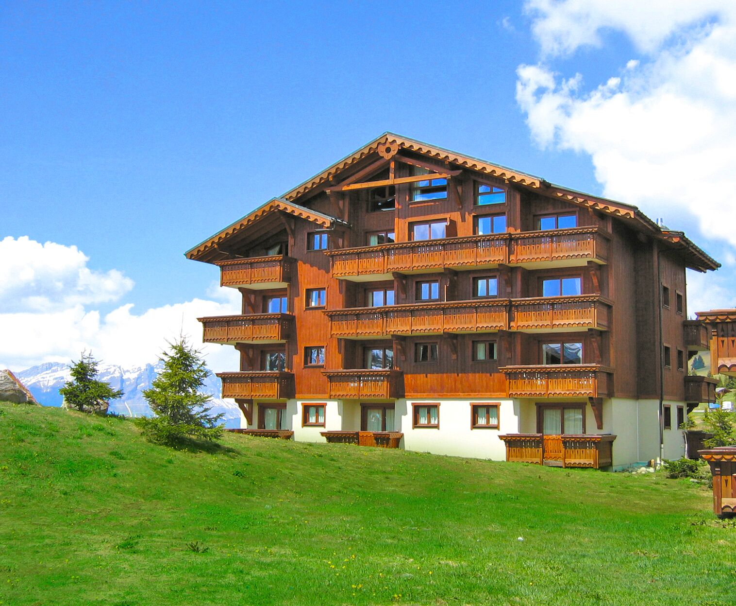 Un bâtiment en bois à plusieurs étages, doté de balcons, situé sur une colline verdoyante avec une chaîne de montagnes en arrière-plan.