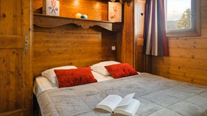 A wooden cabin-style room with a bed, red pillows, a book on the bed, and a shelf with a box and a picture frame. - Residence Lagrange Vacances le Village des Lapons (Hauteluce)