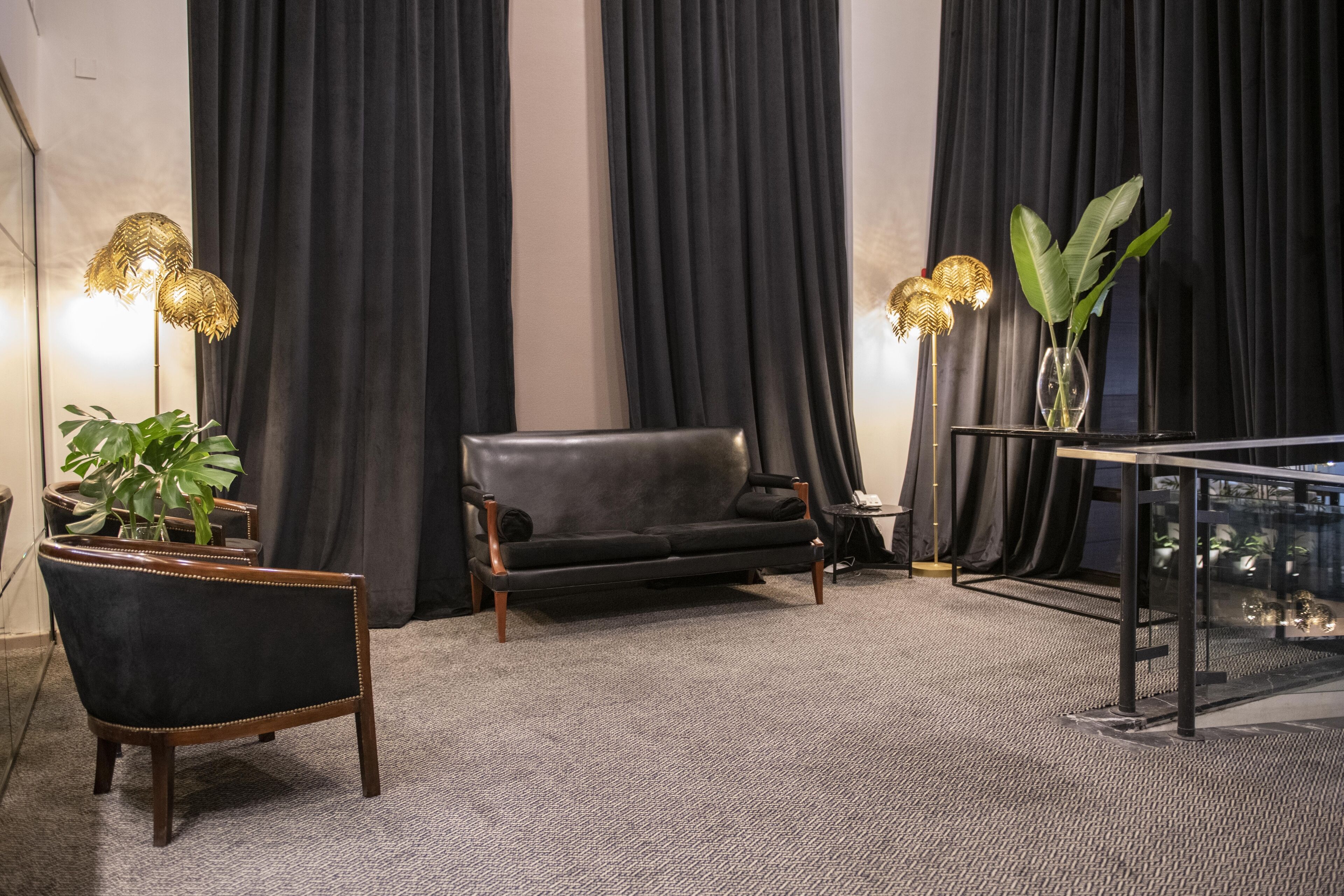 A modern living room with a dark leather sofa, two gold lamps, a glass coffee table, and a large plant.