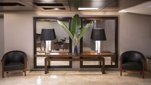 A lobby with a wooden bench, two black leather chairs, a large mirror, and a potted plant. - Grand King Hotel (Buenos Aires)