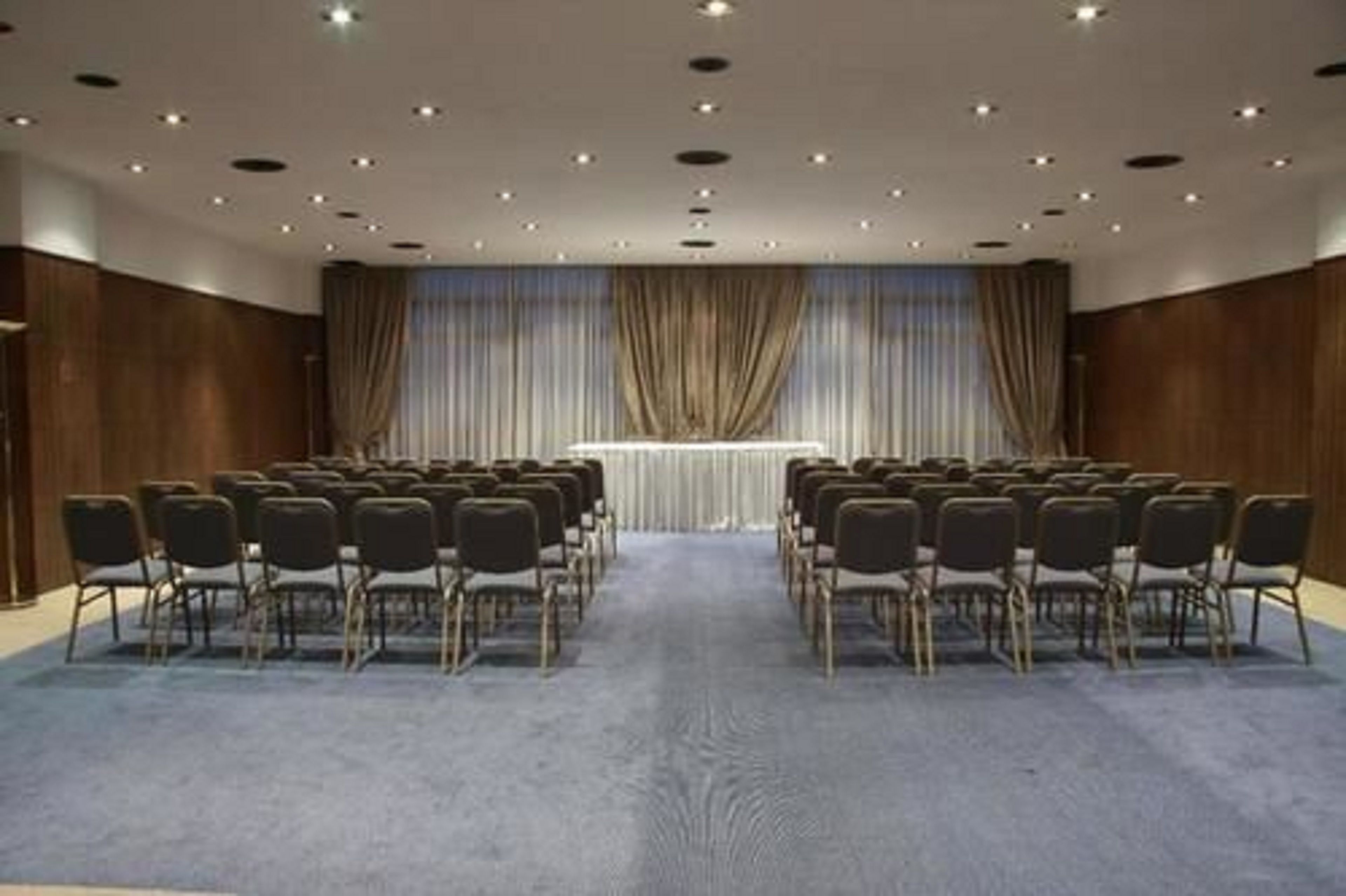 A conference room with rows of chairs facing a stage, wooden paneling on the walls, and a carpeted floor.