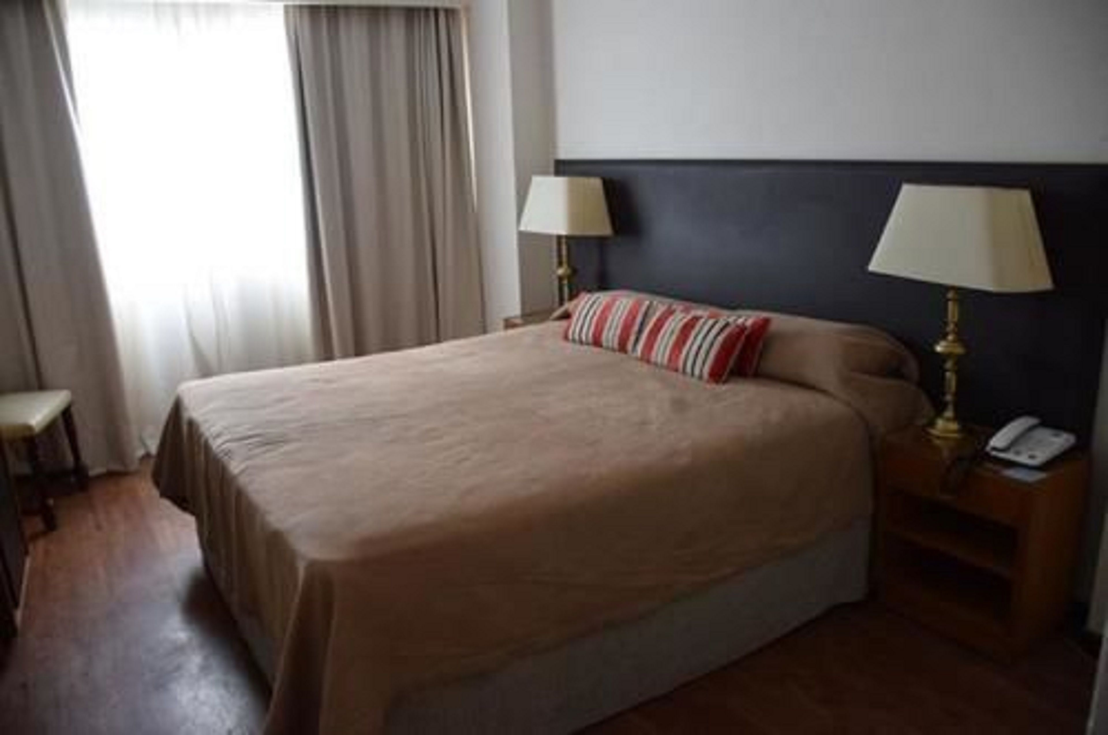 A hotel room with a bed, two bedside lamps, a nightstand, a chair, and a window with curtains.