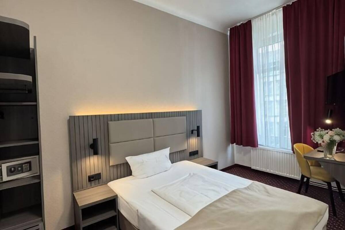 Habitació d'hotel amb llit, taules de nit, escriptori, cadira i una finestra gran amb cortines.
