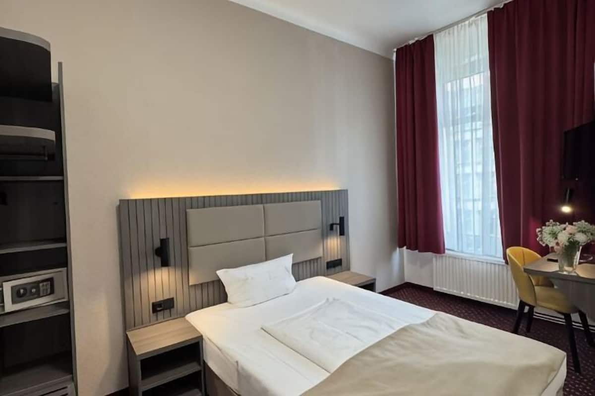 A hotel room with a bed, bedside tables, a desk, a chair, and a large window with curtains.
