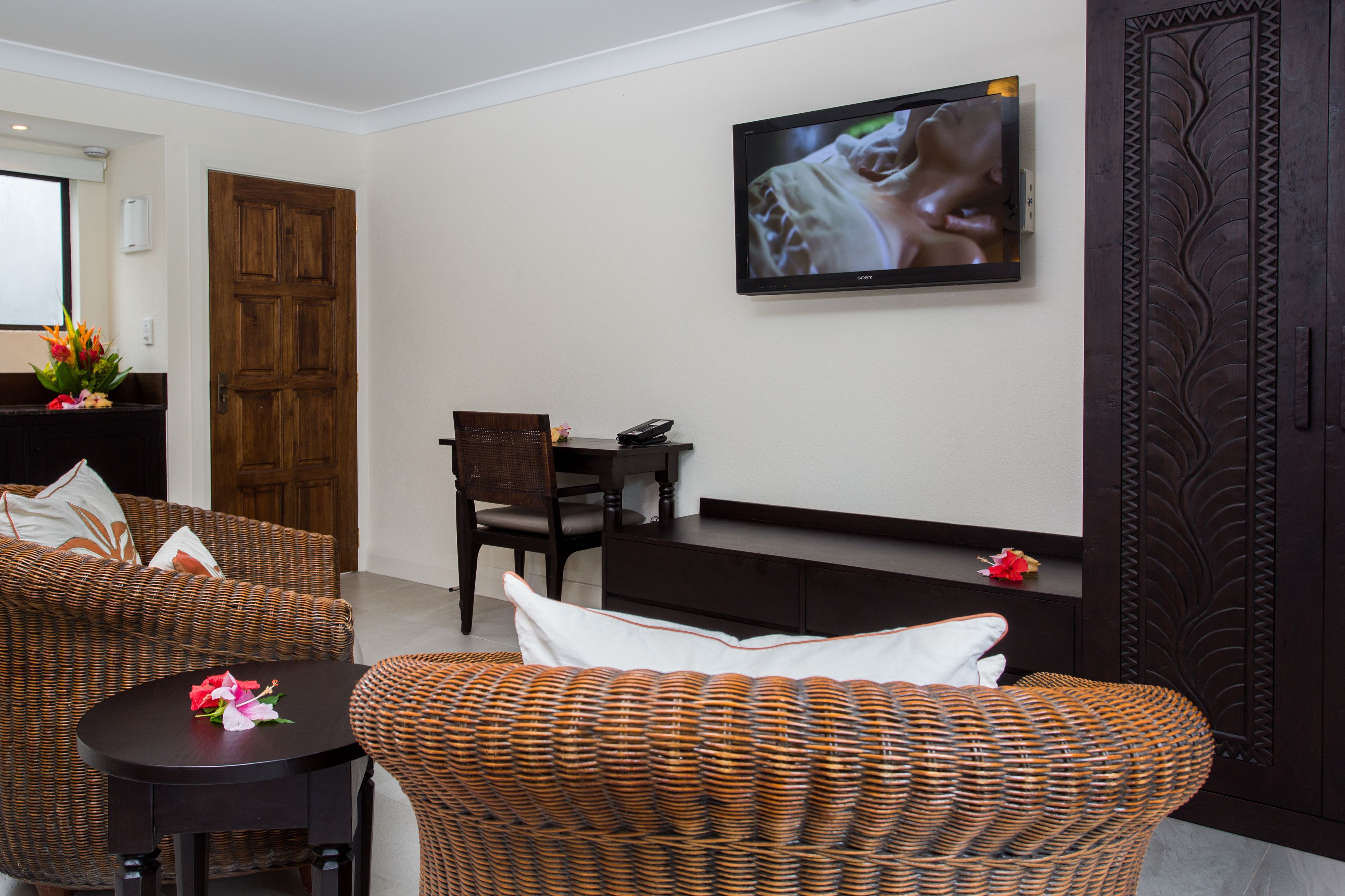 A living room with wicker furniture, a flat-screen TV mounted on the wall, and a wooden door.