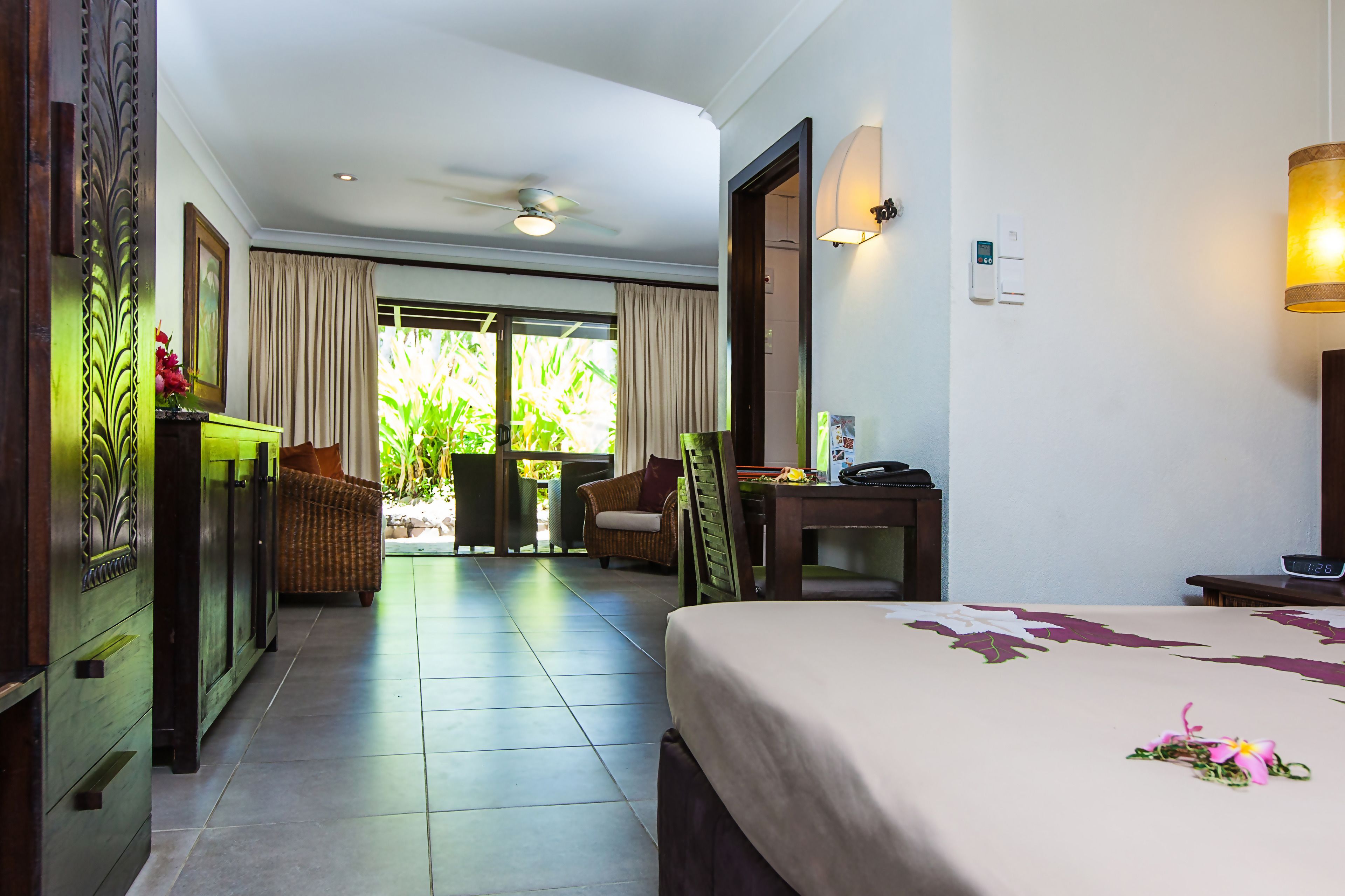 A hotel room with a bed, a desk, a chair, a small table, and a view of greenery outside.