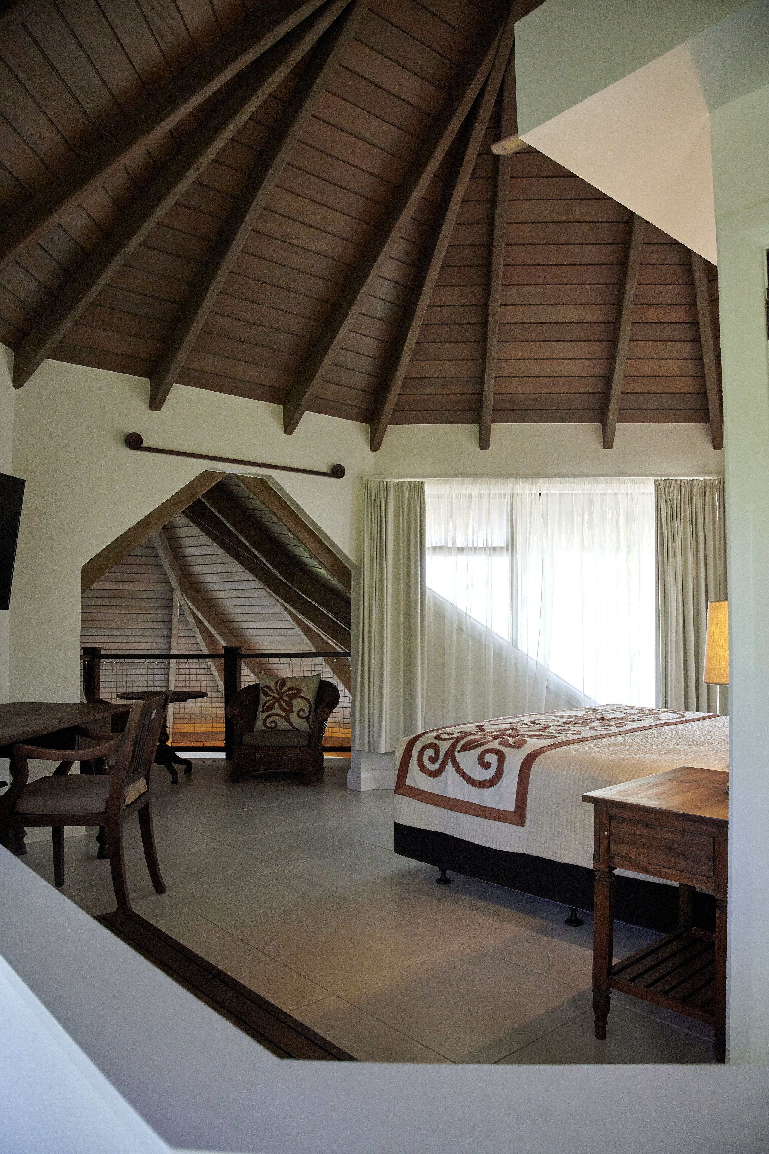 A bedroom with a sloped ceiling, a bed, a desk, a chair, and a window with curtains.