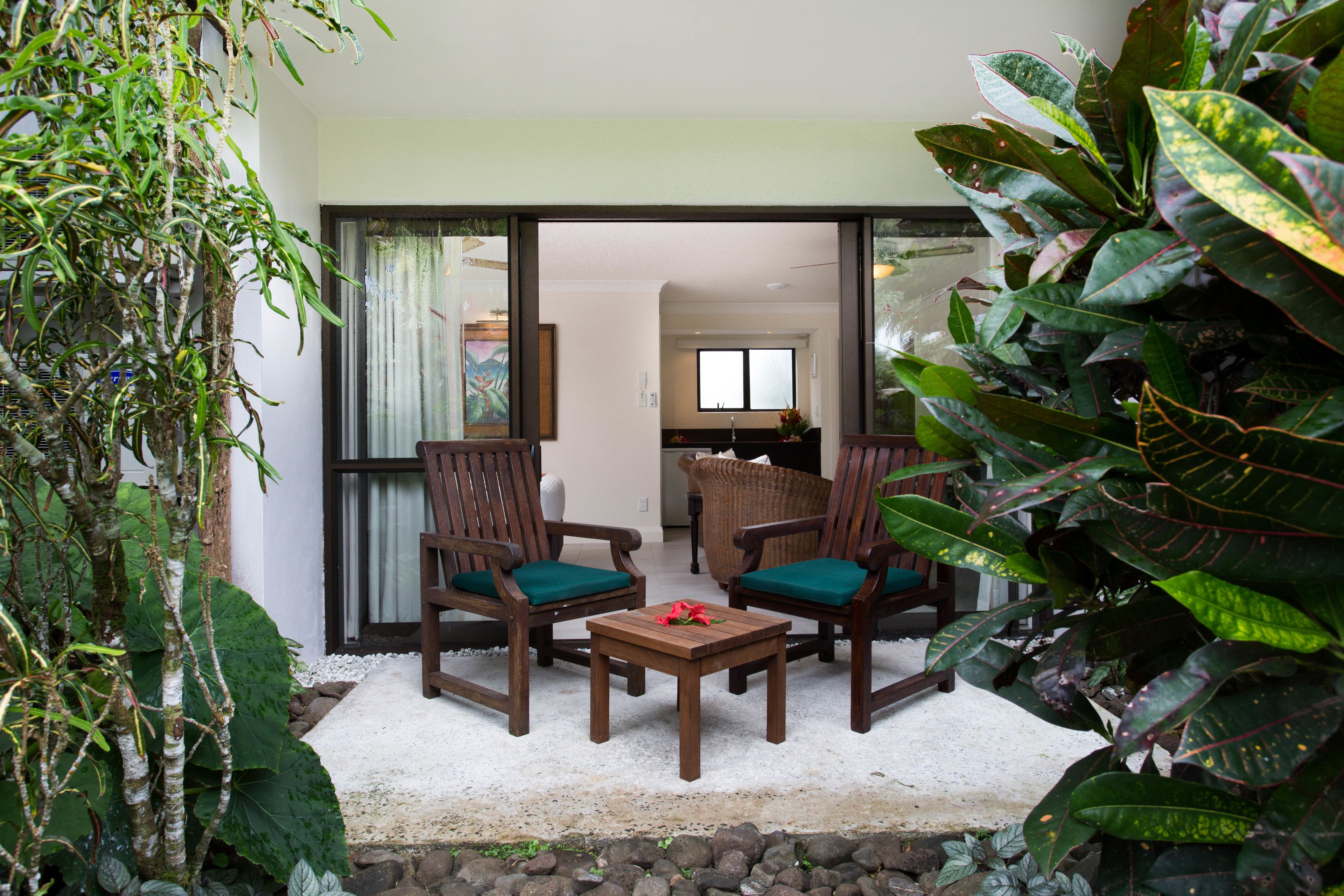 Un patio con una mesa y dos sillas, rodeado de plantas grandes y vistas al interior.