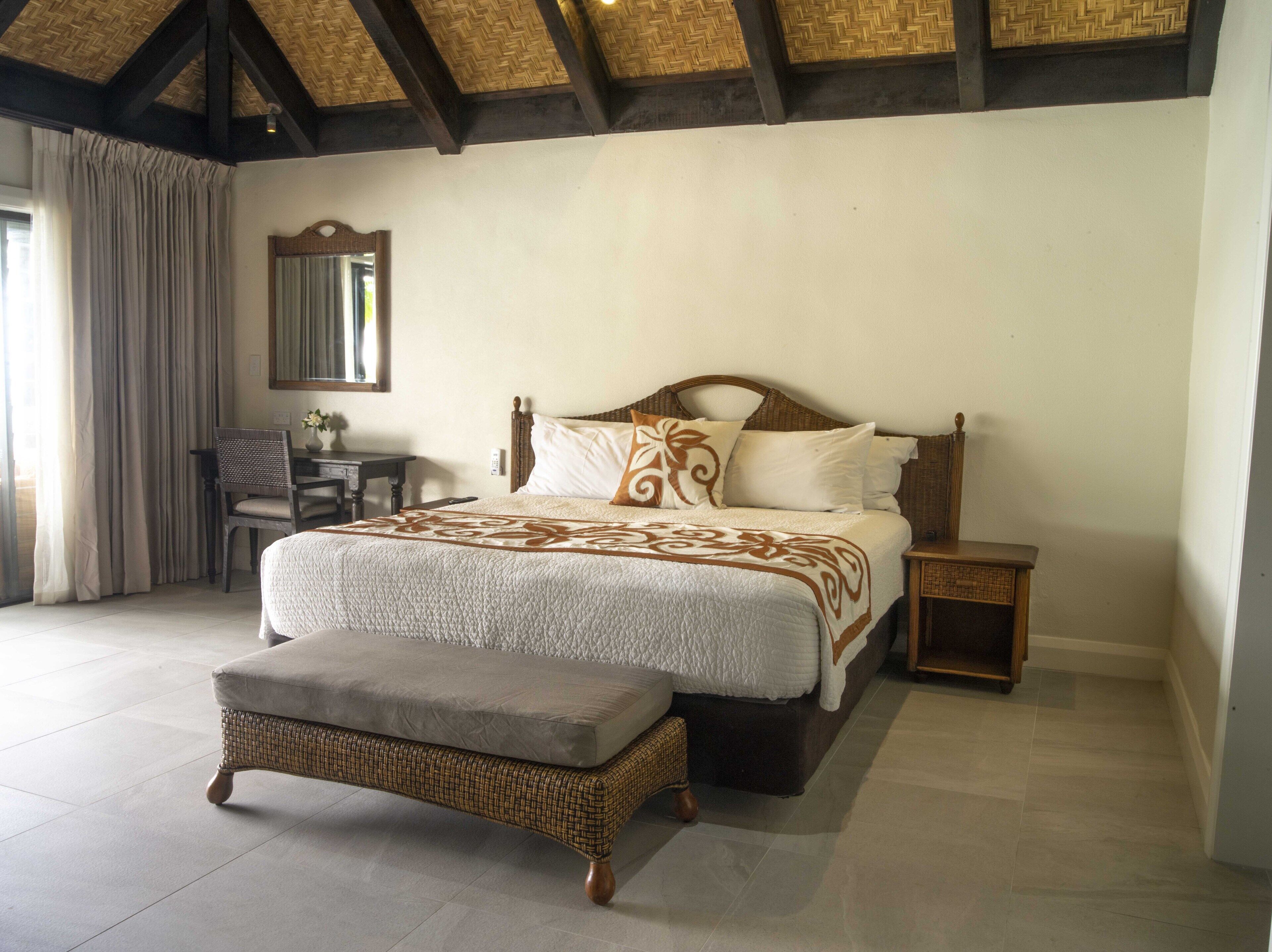 A bedroom with a large bed, a nightstand, a wicker bench, a desk, and a mirror.