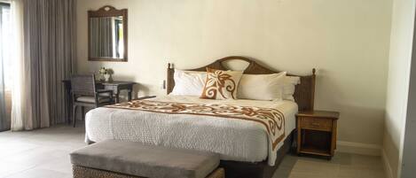 A bedroom with a large bed, a nightstand, a wicker bench, a desk, and a mirror.
