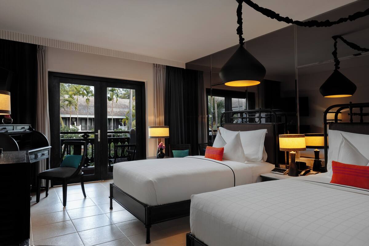 A hotel room with two beds, a desk, and a chair. There are curtains, a lamp, and a view of the outside.