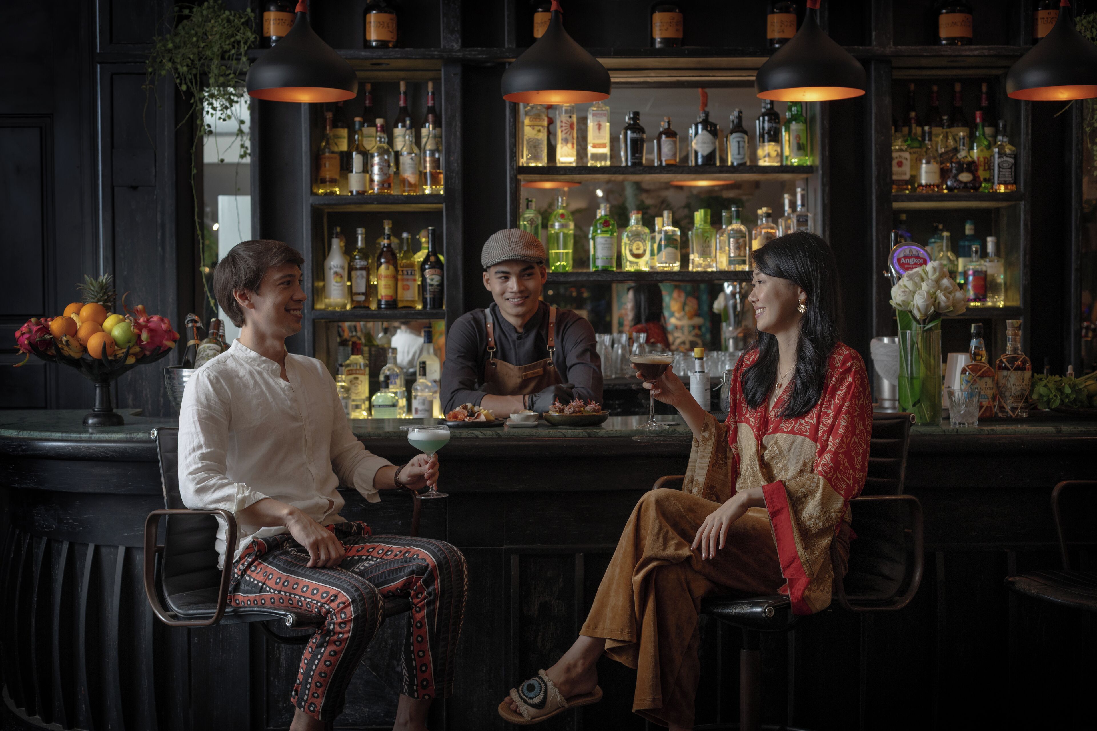 Un bar con tres personas sentadas, diversas botellas en estanterías y un frutero sobre la barra.