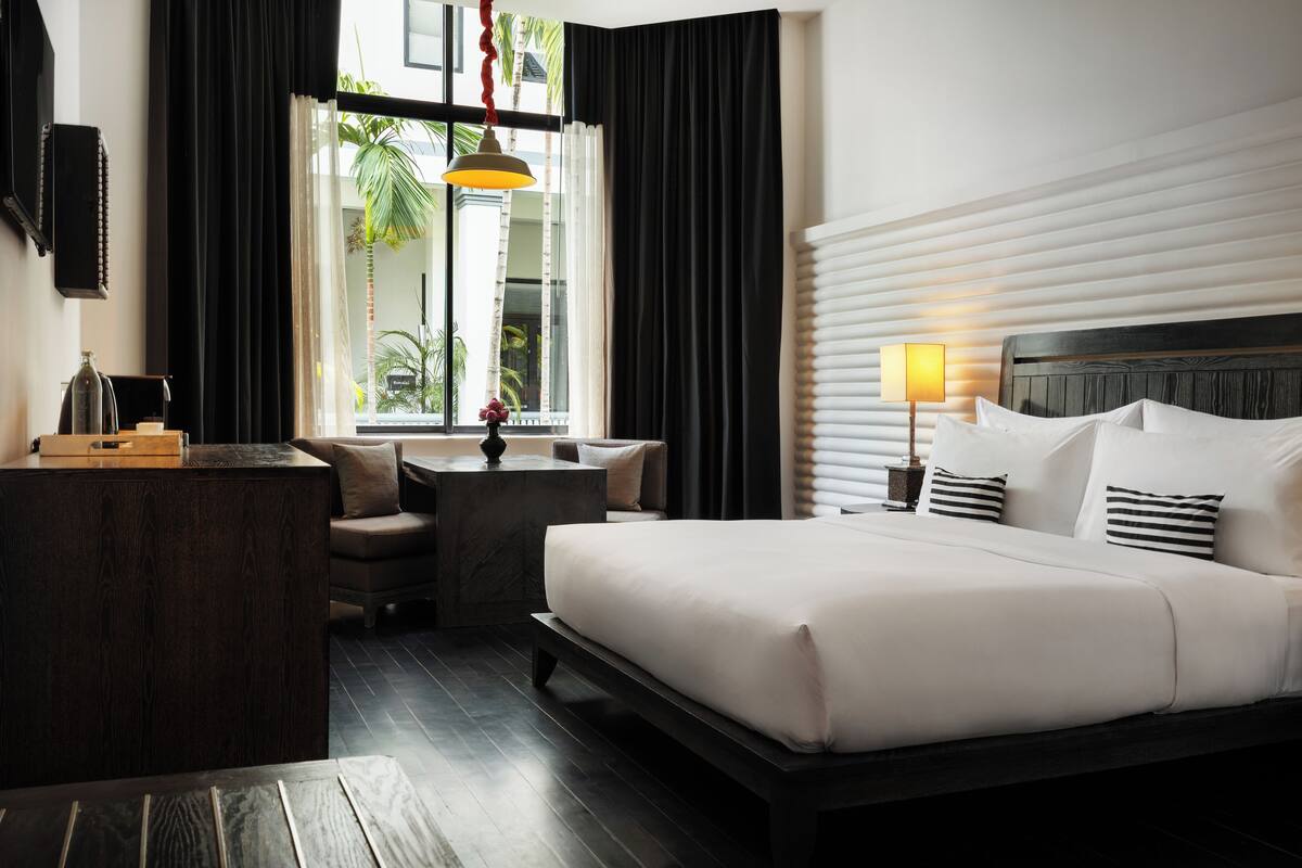 A hotel room with a bed, bedside table, lamp, and a view of the outdoors.