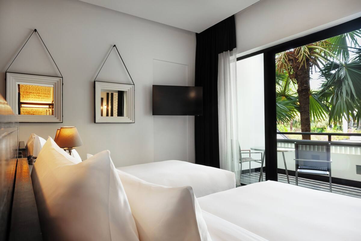 A hotel room with two beds, a flat-screen TV, a window with a view of palm trees, and a balcony with a chair.