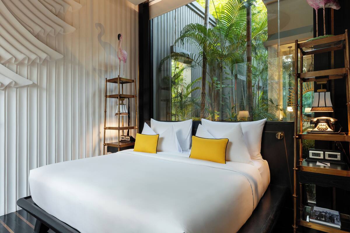 A large bed with white linens and two yellow pillows, a nightstand with a lamp, and a view of greenery through a window.