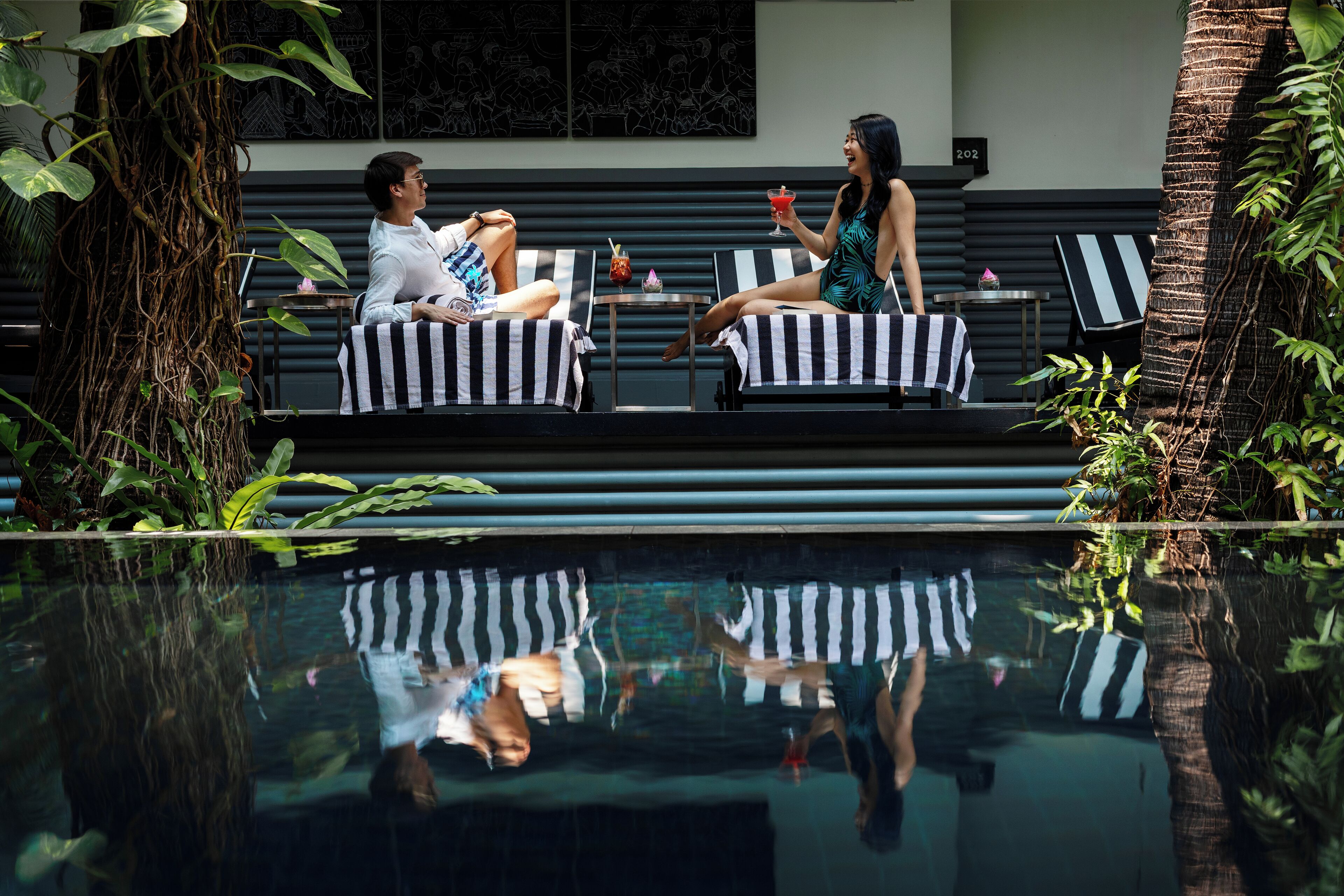 Dos personas sentadas en sillones a rayas junto a una piscina, con follaje tropical de fondo.