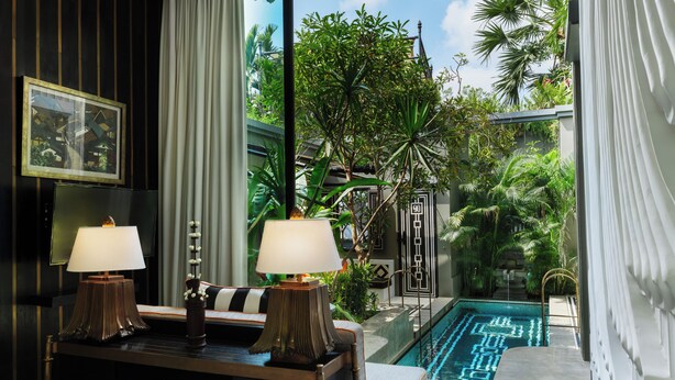 A hotel room with a bed, two bedside lamps, a view of an indoor swimming pool, and lush greenery outside.