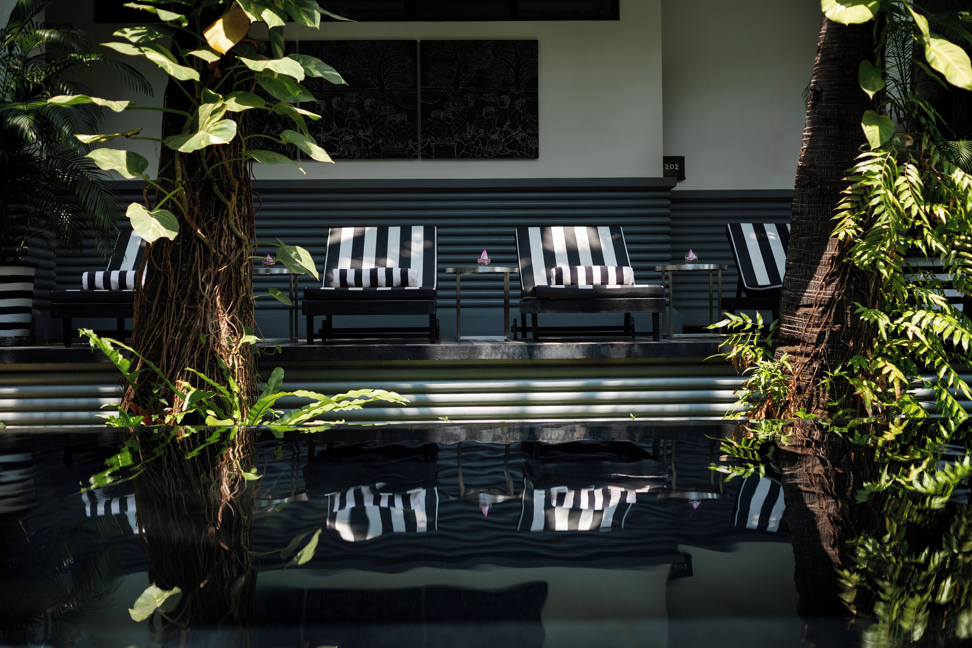 A modern outdoor seating area with striped lounge chairs and a small table, surrounded by lush greenery and a reflective pool.