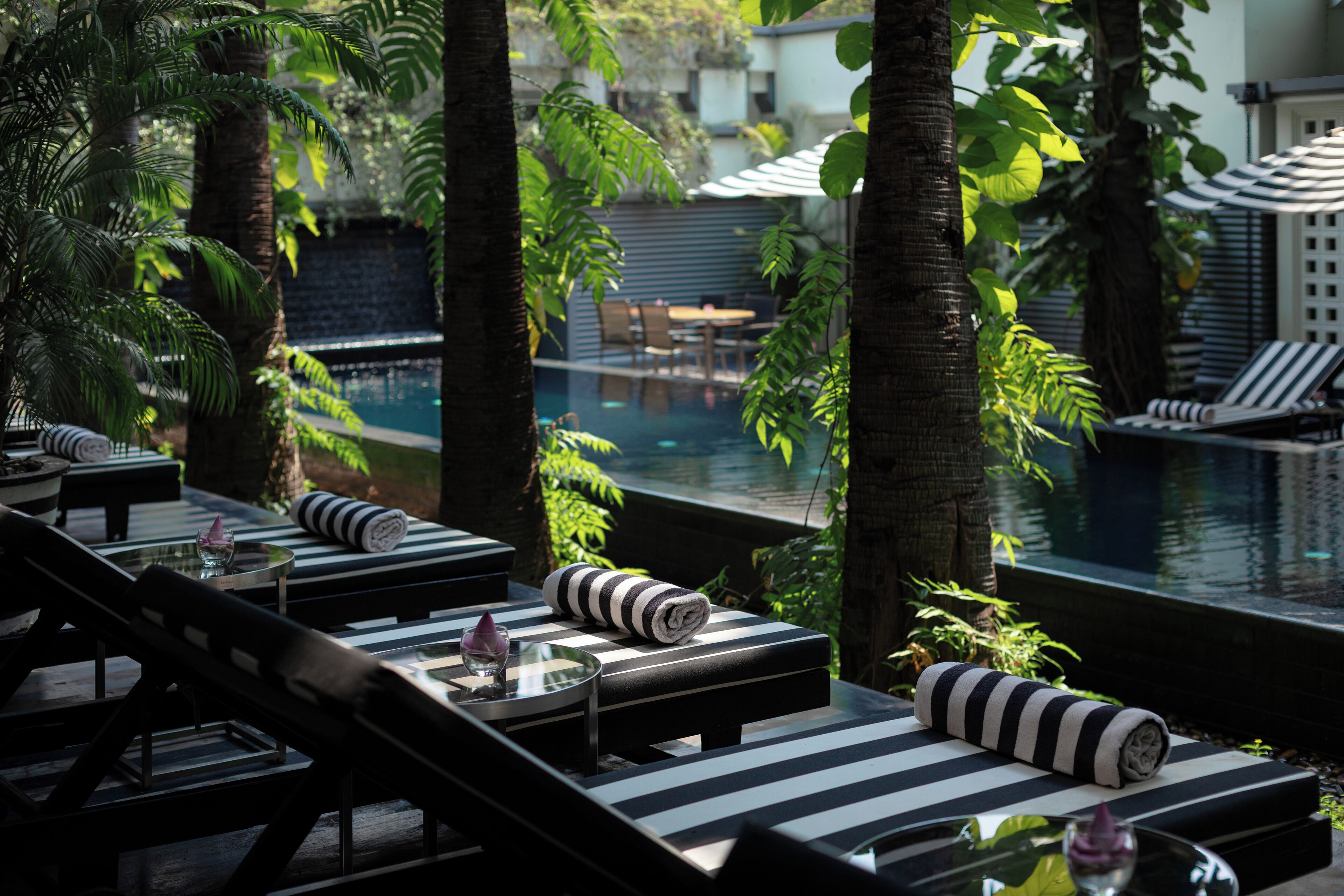 A poolside area with lounge chairs and a table, surrounded by palm trees and a view of a swimming pool.