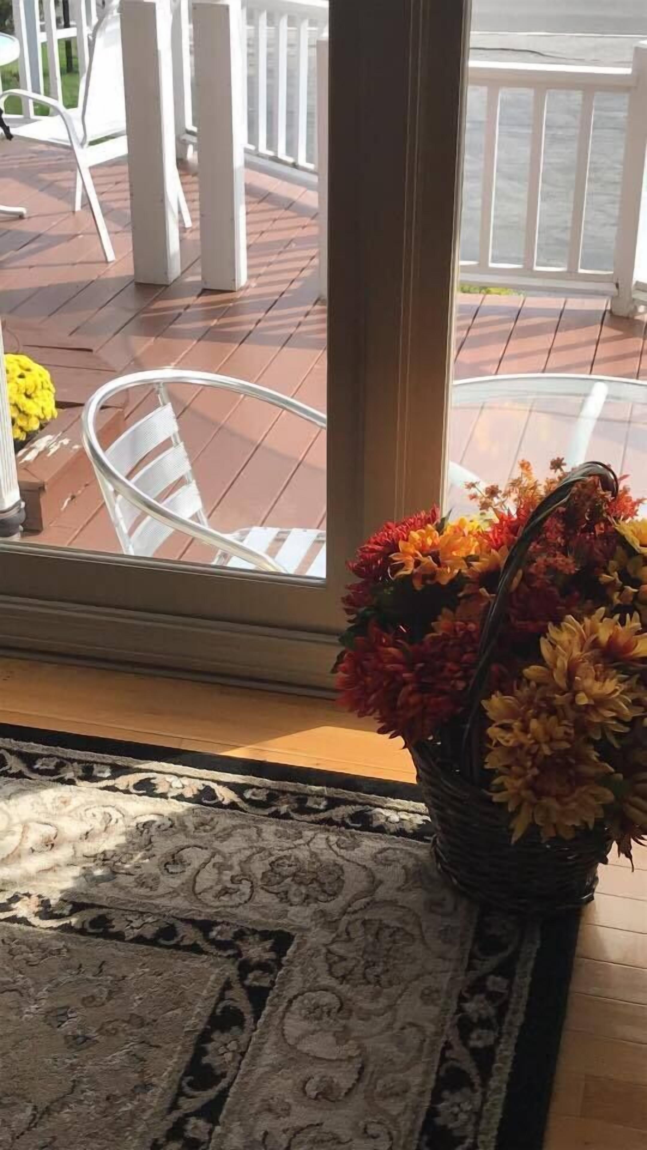 A view through a window to an outdoor deck with a white railing, a wicker chair, and a floral arrangement on a rug.