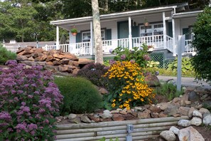 Una casa con un porche, rodeada por un jardín con diversas plantas y un montículo de rocas.