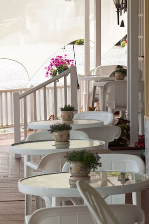 Un área de estar al aire libre techada, con sillas y mesas de plástico blanco, decorada con plantas y flores en macetas.