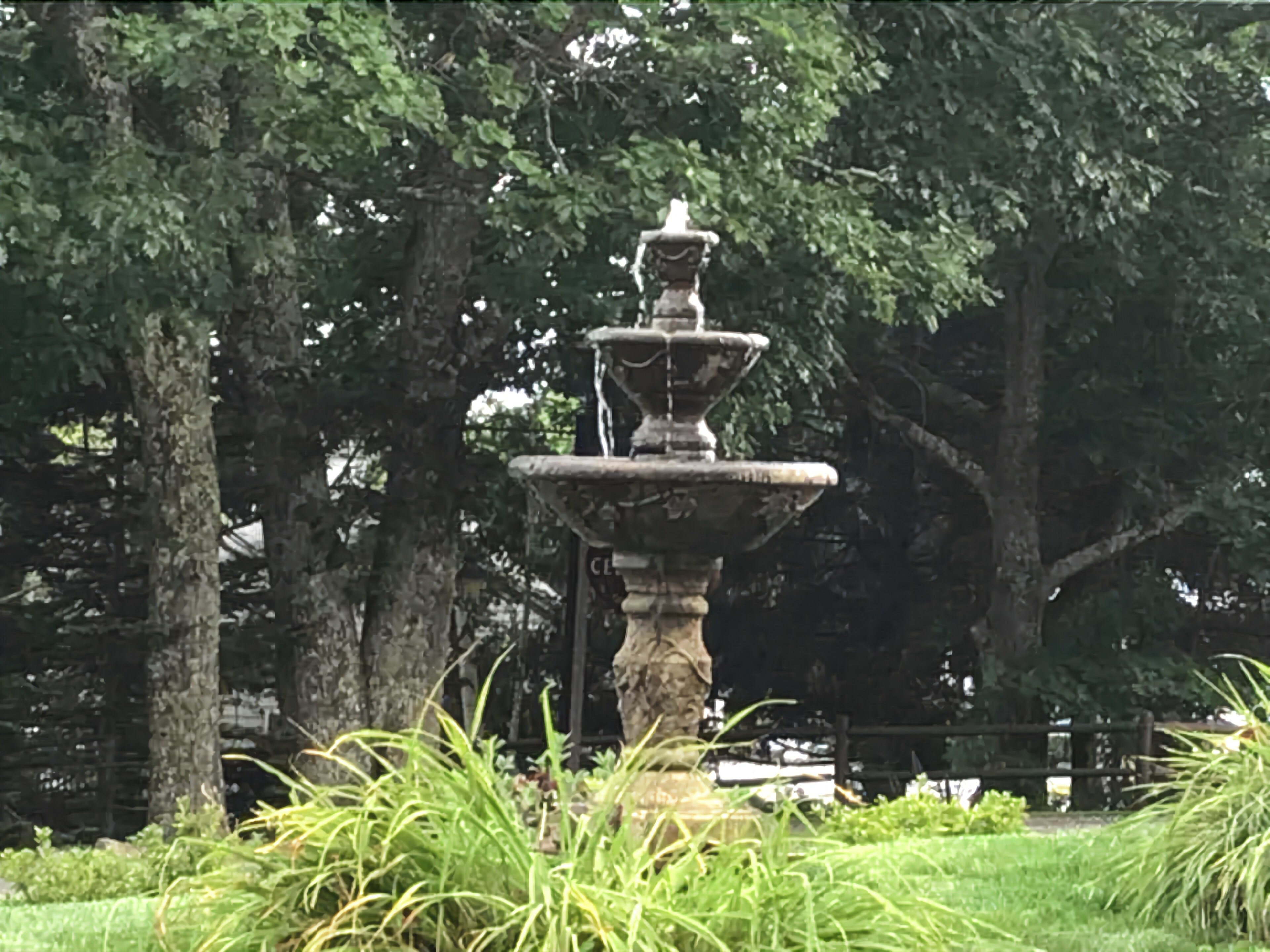 Een stenen fontein met meerdere lagen, omringd door weelderige begroeiing en bomen.